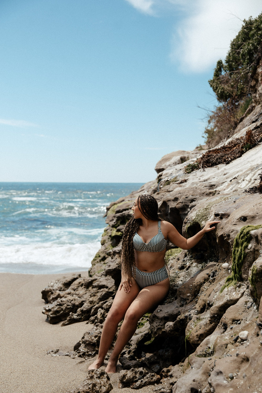 A woman model with long hair sits on rocky cliffs overlooking the ocean in Laguna, wearing a navy blue and white striped bikini that provides ample seat coverage with higher cut bottoms.